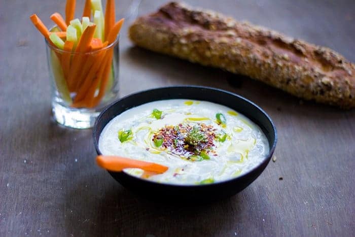 A bowl of Garlicky Yogurt Dip with a glass of long slit veggies and a loaf of bread
