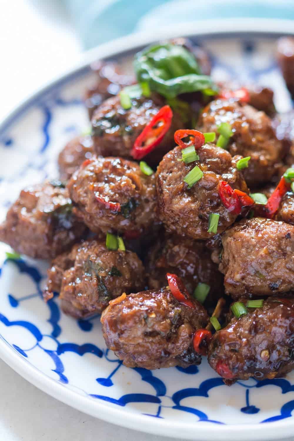 close up shot of sticky meatballs garnished with spring onions and thai red chillies served in a white and blue plate