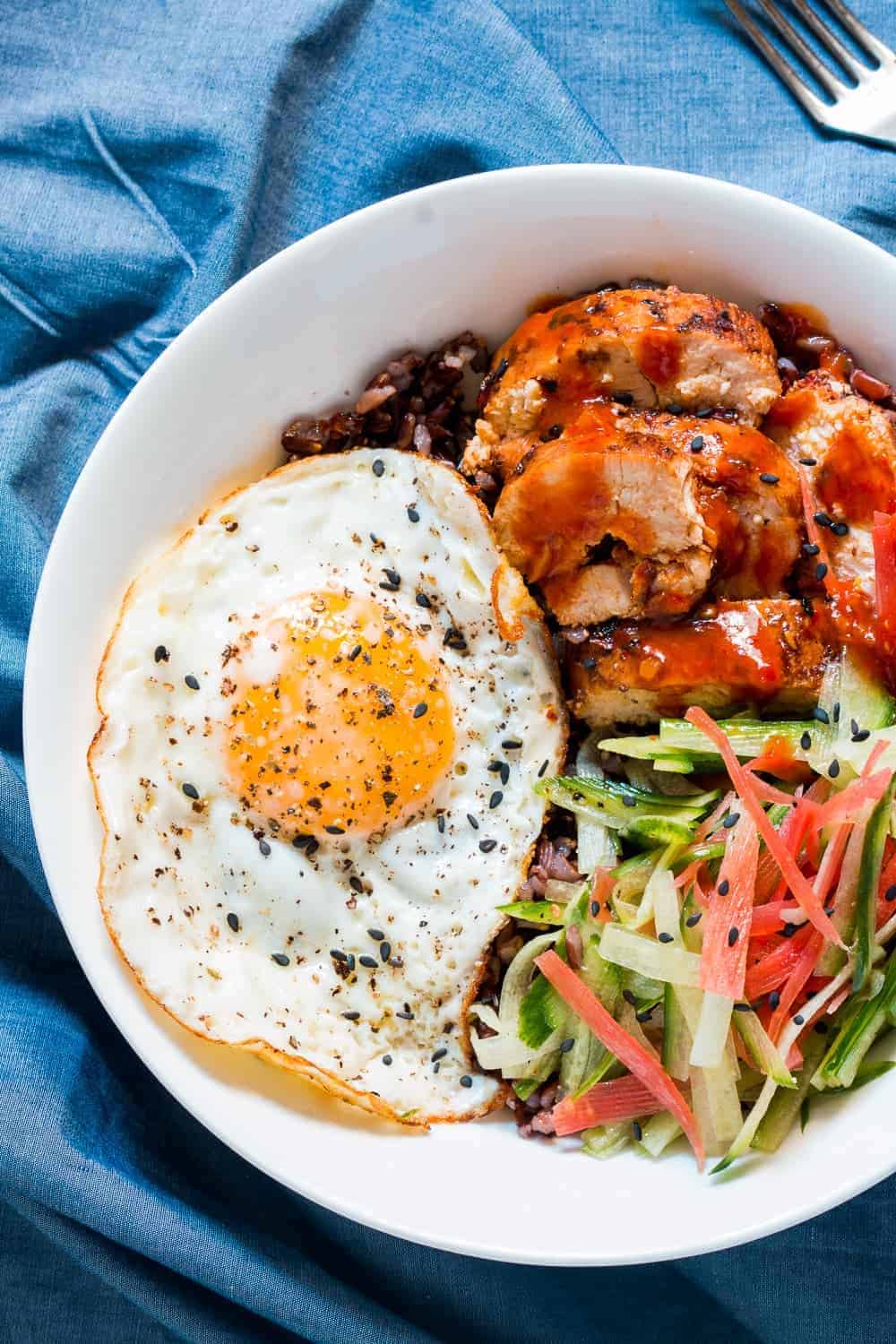 a white bowl of schezwan chicken, sliced veggies, black rice and fried egg