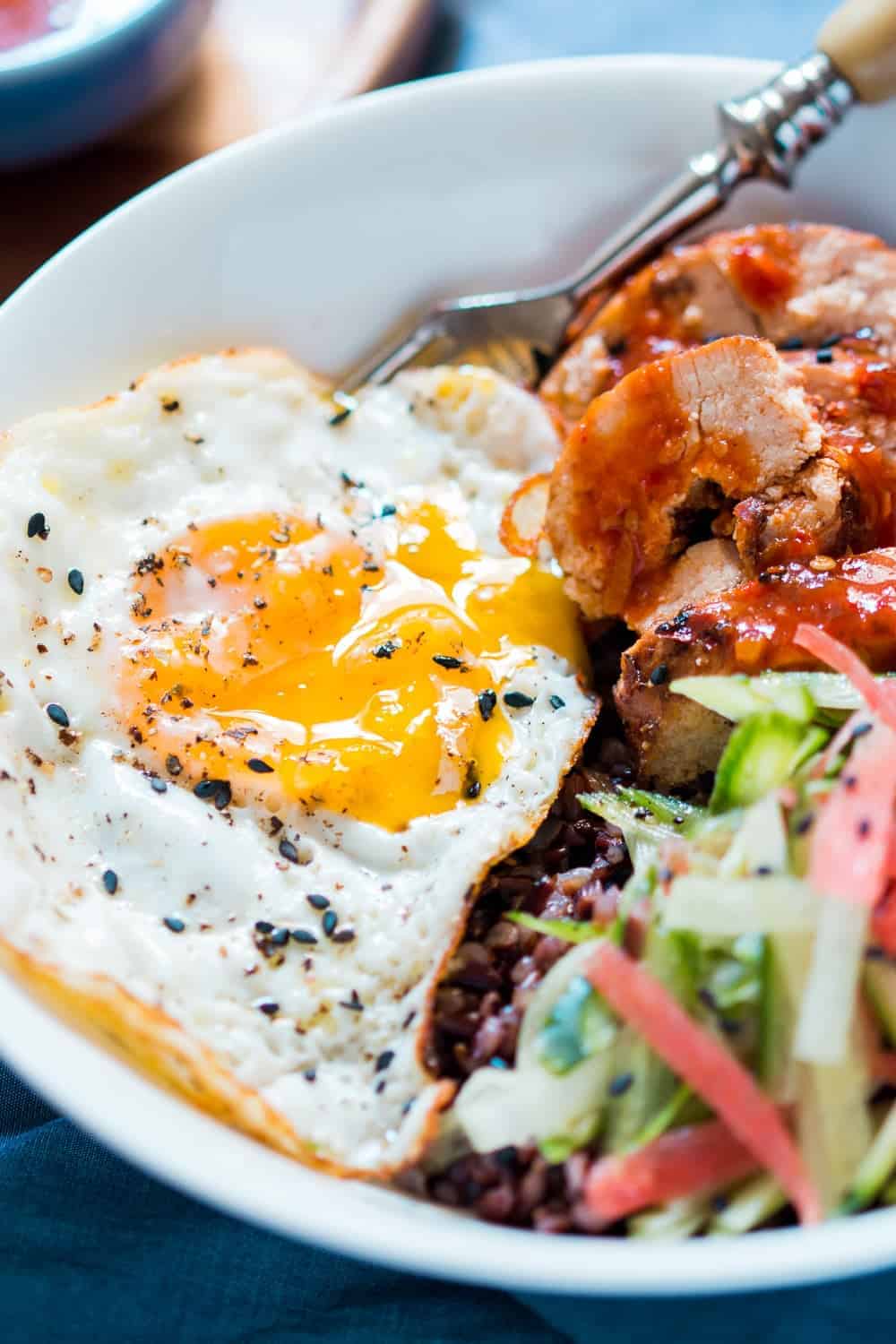 close up of a white bowl of schezwan chicken, sliced veggies, black rice and fried egg