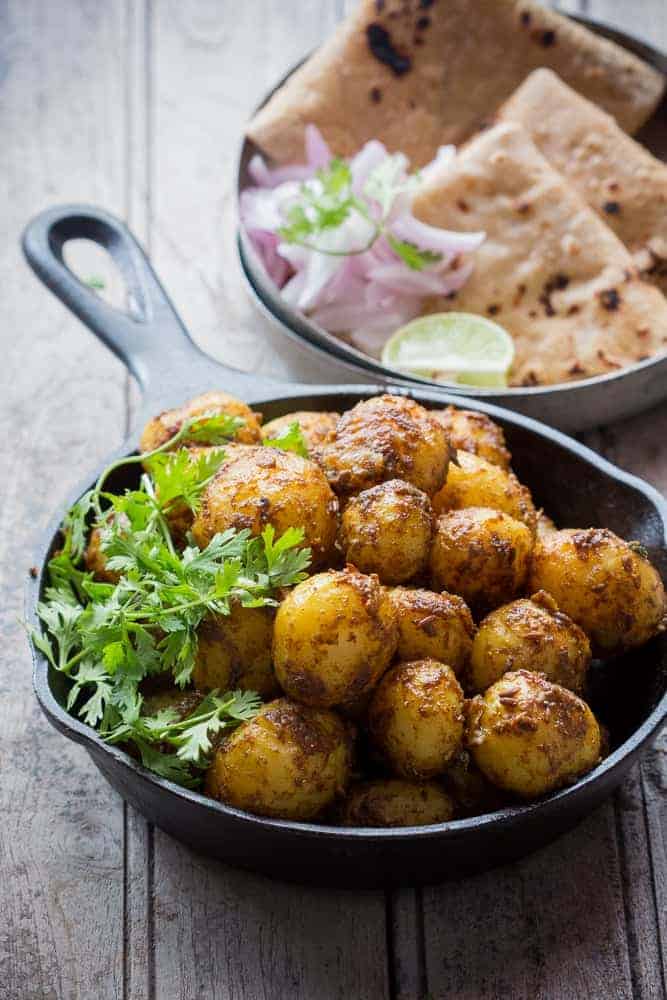 shot of bombay potatoes with coriander in a cast iron pan with onions, lemon and rotis in a plate on the side