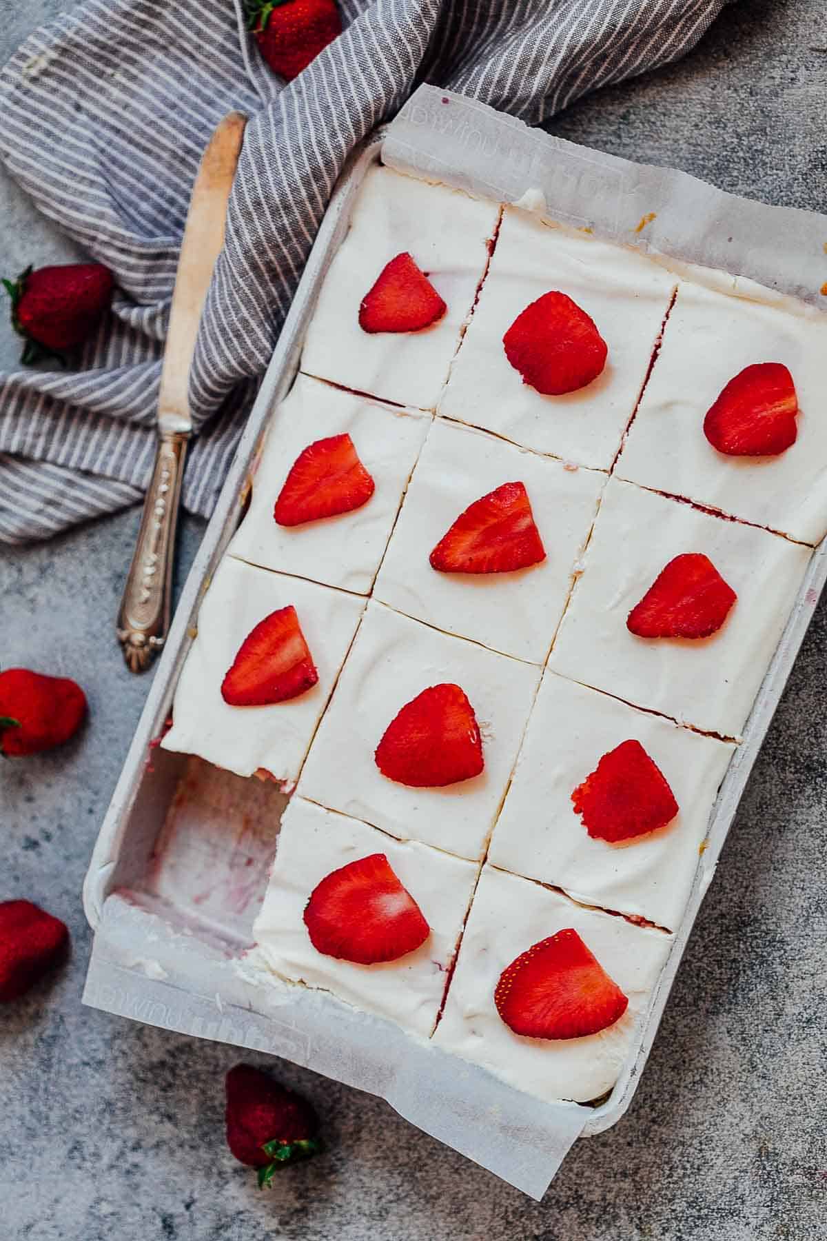 Slices of strawberry poke cake in a sheet pan with a knife and fresh strawberries on the side