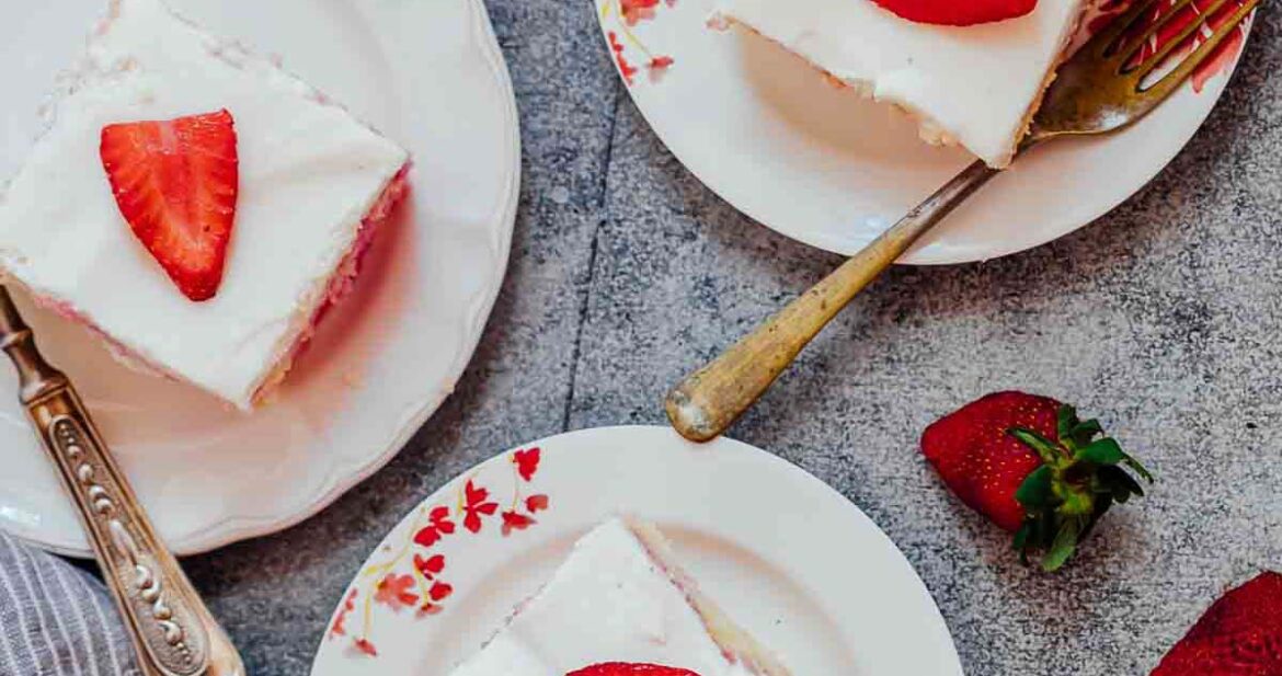 Three slices of strawberry poke cake made from scratch served on white plates with forks and fresh strawberries on the side