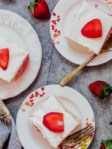 Three slices of strawberry poke cake made from scratch served on white plates with forks and fresh strawberries on the side