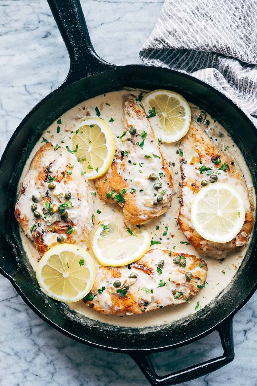 Creamy lemon chicken scallopini served in a cast iron skillet with four chicken breasts with capers and lemons on top