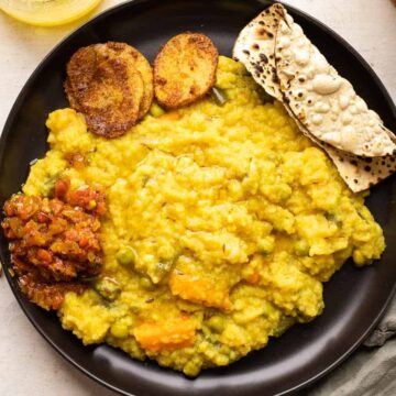 Khichdi served on a black plate with papad, tomato chutney and aloo fry