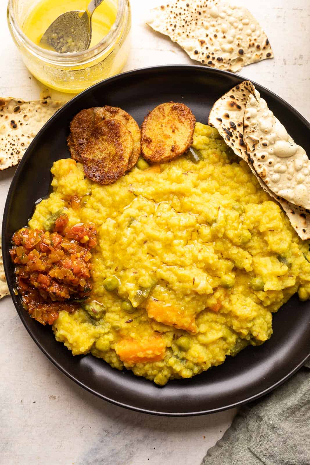 Khichdi served on a black plate with papad, tomato chutney and aloo fry