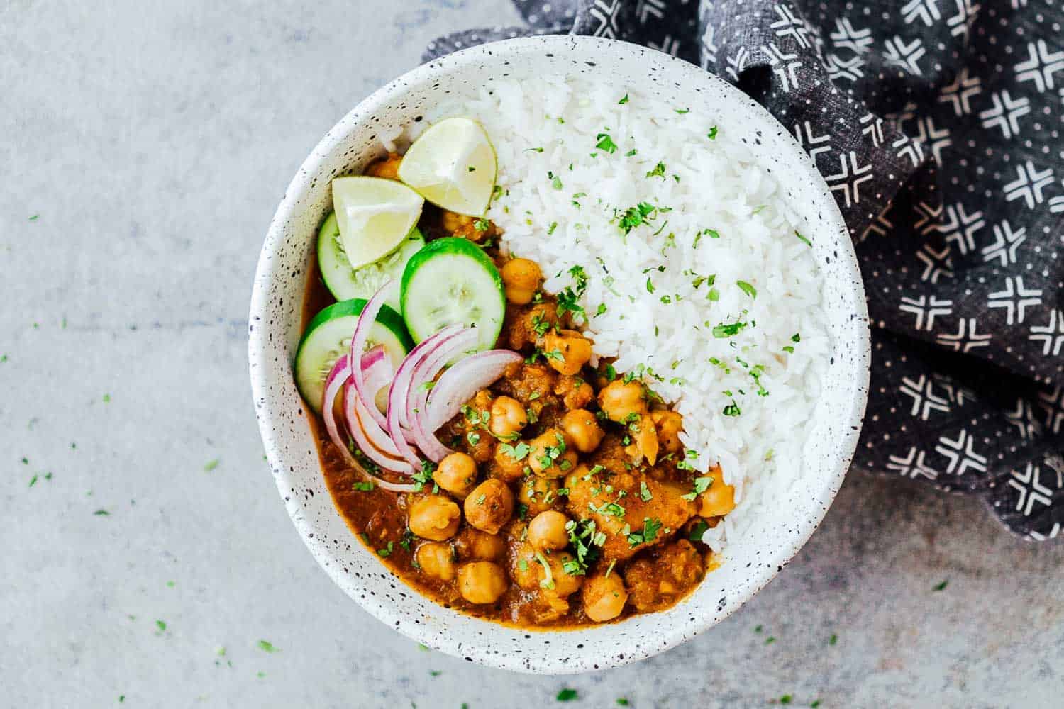 A bowl of chana masala, rice with sliced onions. cucumbers and lemons on the side