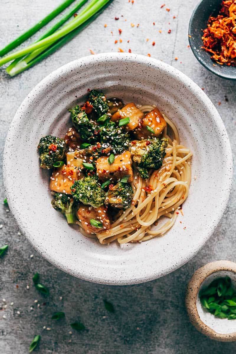 Crispy tofu broccoli stir fry in a white bowl topped with chilli flakes, green onions and sesame seeds