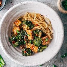 Crispy tofu broccoli stir fry in a white bowl topped with chilli flakes, green onions and sesame seeds.