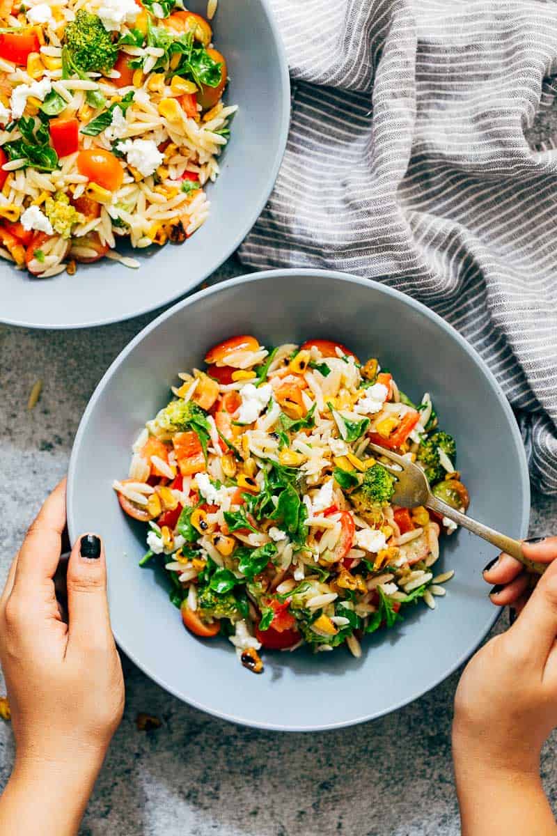 Two bowls of colourful, zippy, summery lemon orzo pasta salad with a hand holding one of the bowls