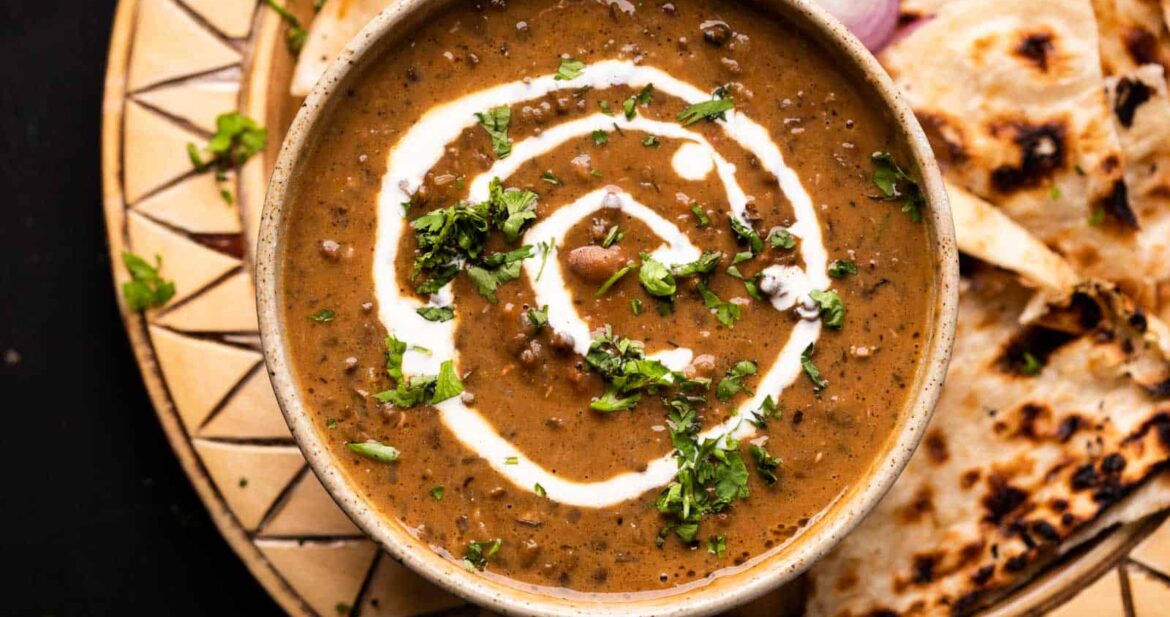 Dal Makhani served in a brown ceramic bowl with cream drizzled on top and naan and sliced onions on the side