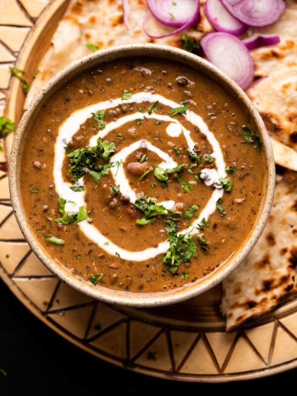 Dal Makhani served in a brown ceramic bowl with cream drizzled on top and naan and sliced onions on the side
