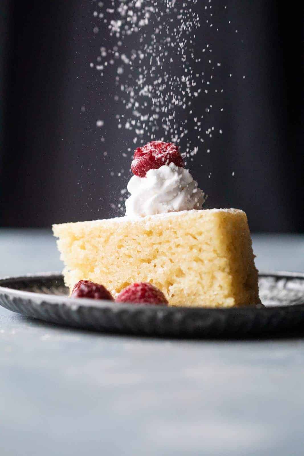 A slice of eggless vanilla cake with whipped cream, raspberries and a sprinkling of icing sugar