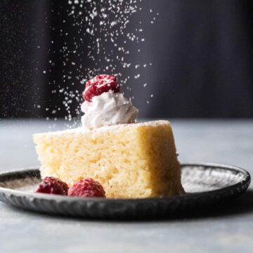 A slice of eggless vanilla cake with whipped cream, raspberries and a sprinkling of icing sugar