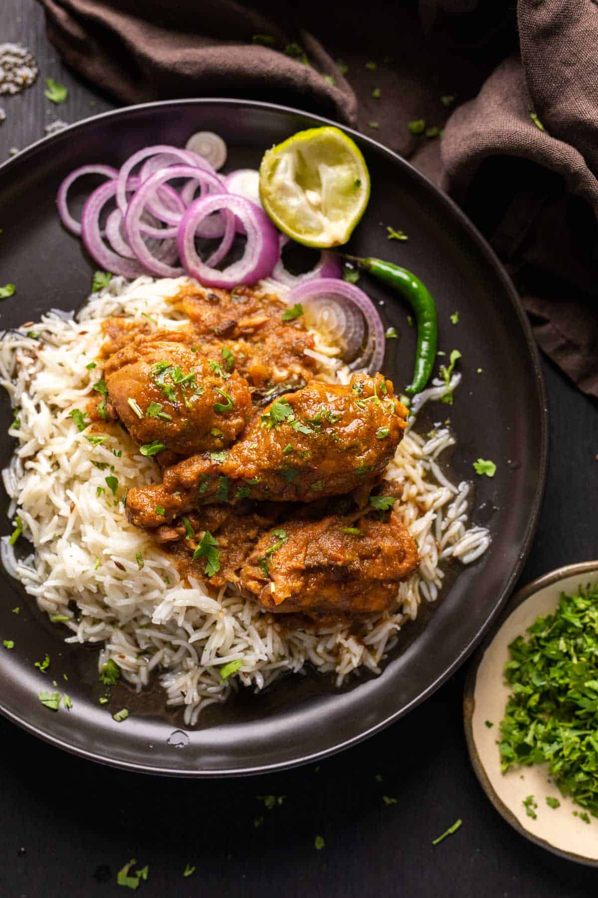 Chicken curry served on a black plate with rice, onions, lime juice and green chillies on the side