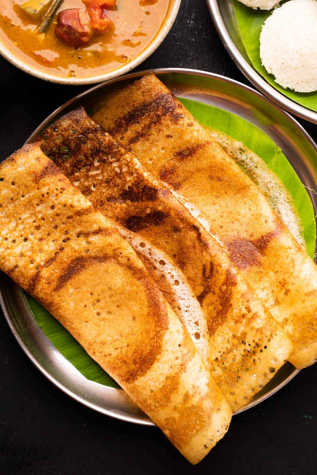 A picture of three crispy dosas laid out on a plate lined with banana leaf with idlis, sambar and chutney on the side