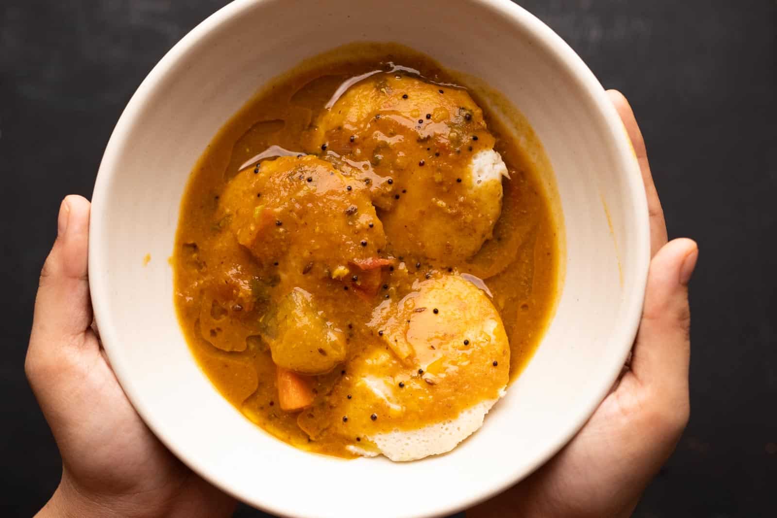Sambar poured over steaming hot idlis in a bowl. Two hands hold the bowl as if serving someone else