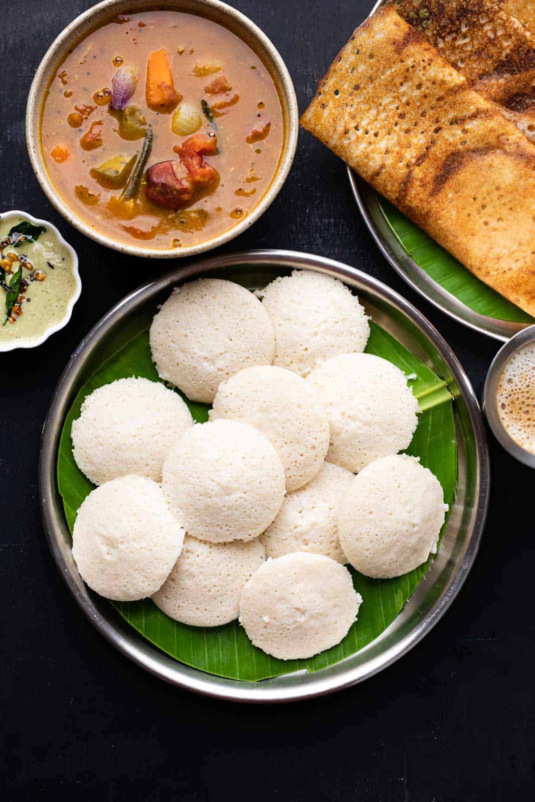 A picture of soft idlis stacked on top of each other on a banana leaf lined plate with dosas, chutney and sambar on the side