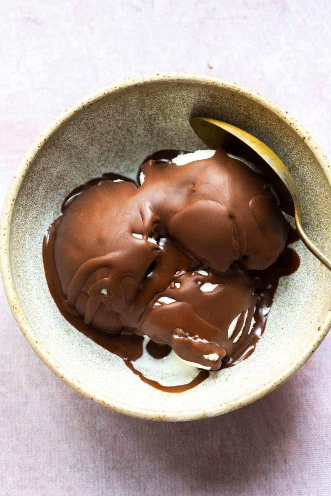 Chocolate magic shell that has hardened over vanilla ice cream to form a crackly crispy shell served in a brown ceramic bowl with a spoon