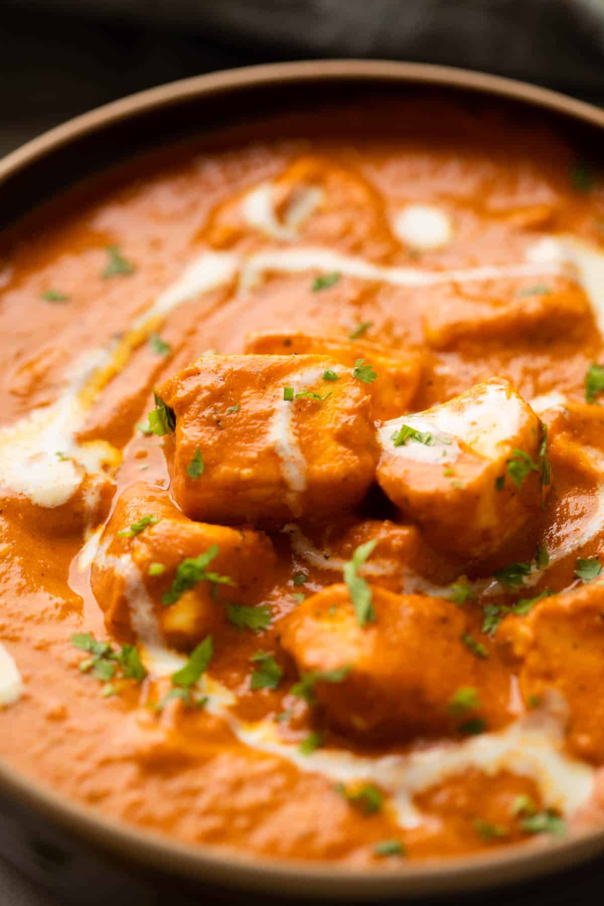 A closeup of paneer pieces in paneer butter masala with dollops of cream and finely chopped coriander on top