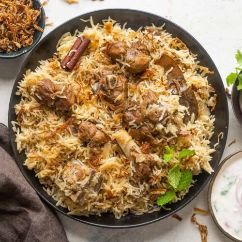 Mutton yakhni pulao served on a black plate with onions and raita on the side