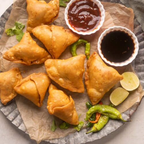 Samosas placed on a plate with ketchup, chutney, fried chillies, chai and lime