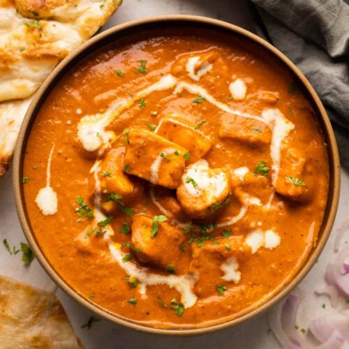 Paneer Butter Masala served in a brown bowl with naan and onions on the side
