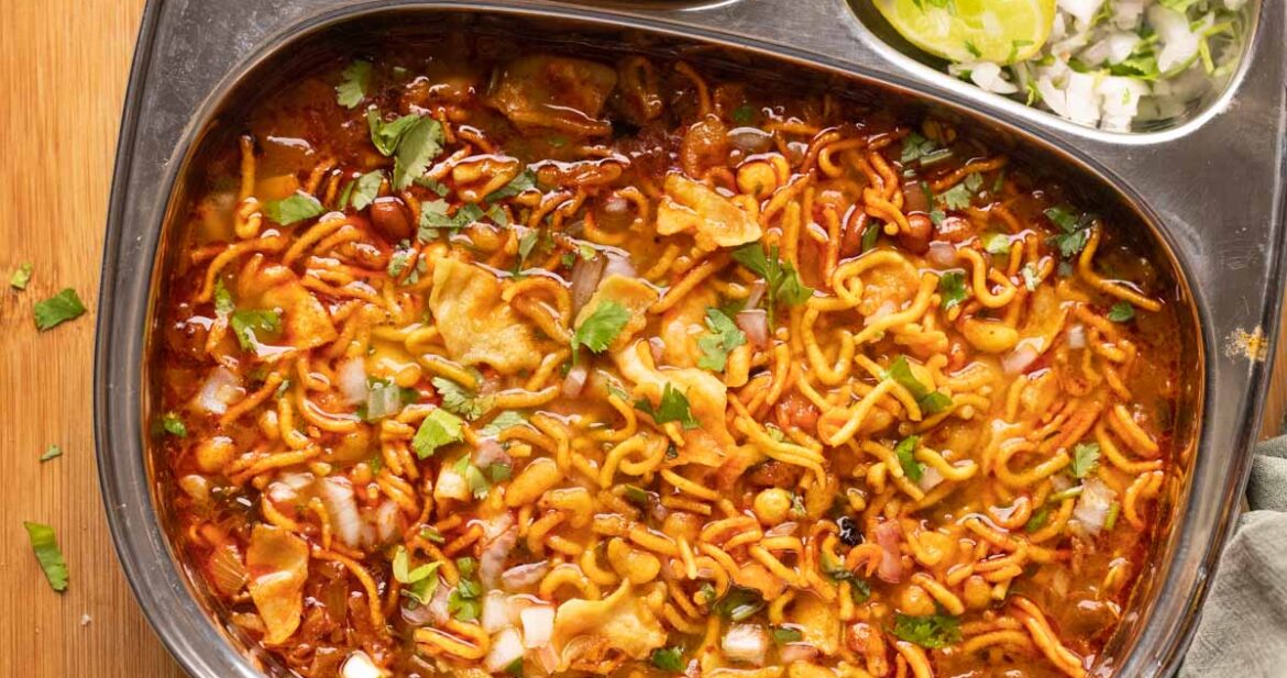 Picture of misal topped with farsaan served in a steel plate with pan, chopped onions and lime wedges on the side