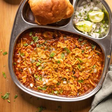 Picture of misal topped with farsaan served in a steel plate with pan, chopped onions and lime wedges on the side