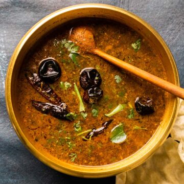 Picture of rasam in the brass pot that it was cooked in with a wooden ladle and some papad on the side