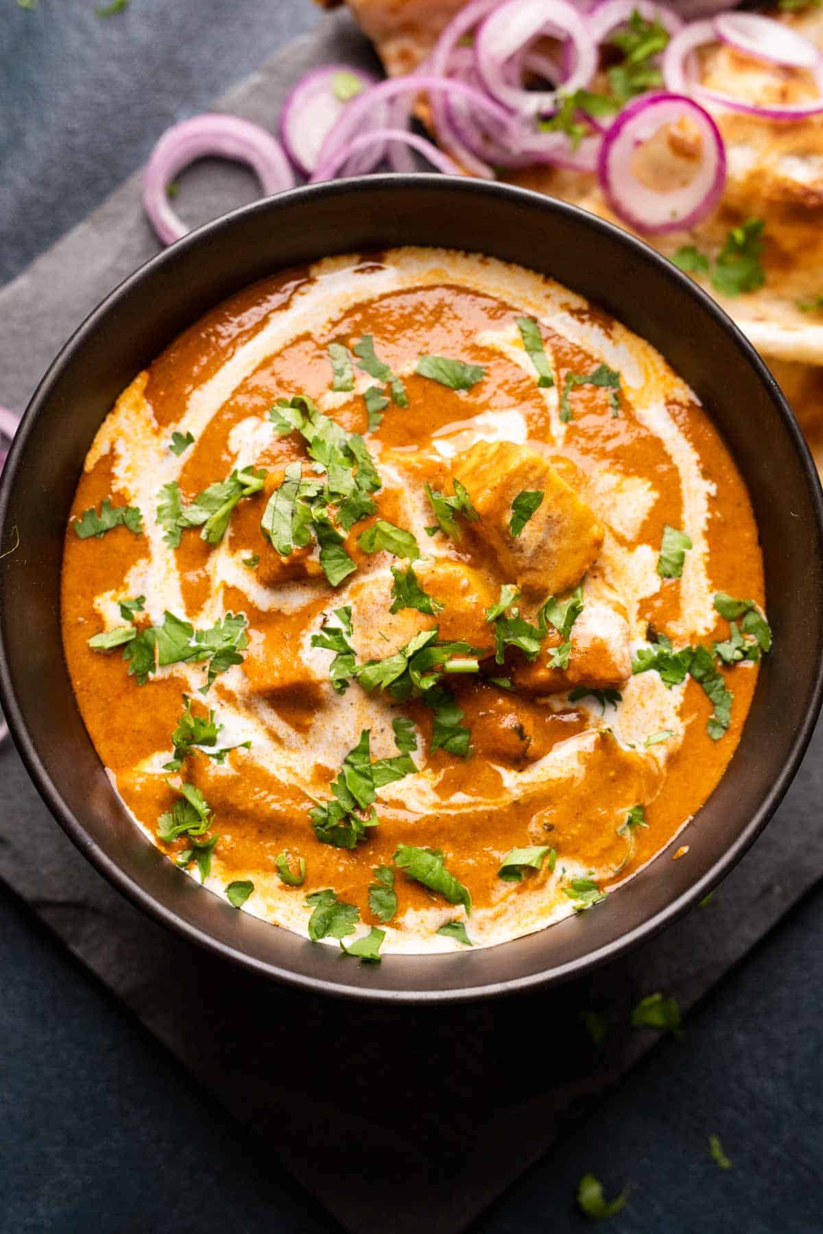 Picture of shahi paneer served in a black bowl with onions and naan on the side