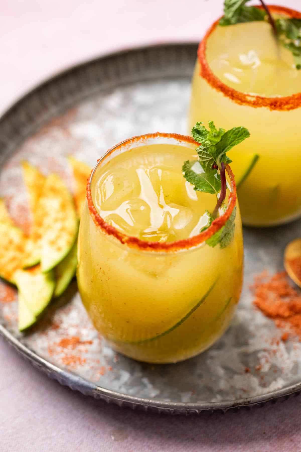 Two glasses of Aam Panna served on a grey platter with sliced raw mangoes and garnishes on the side