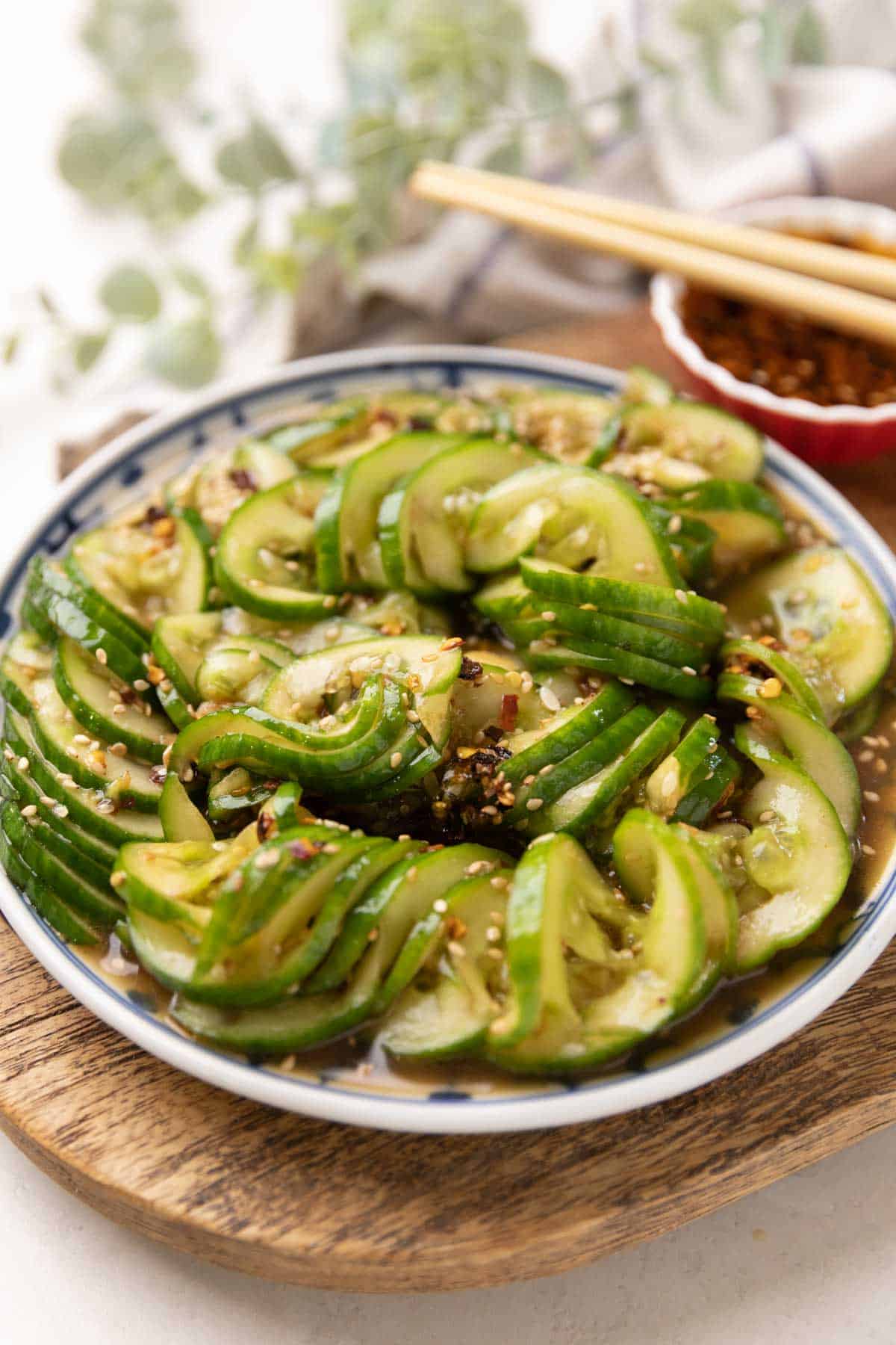 asian cucumber salad served in a blue and white plate placed on a brown board with a red bowl of salad dressing and chopsticks on the side