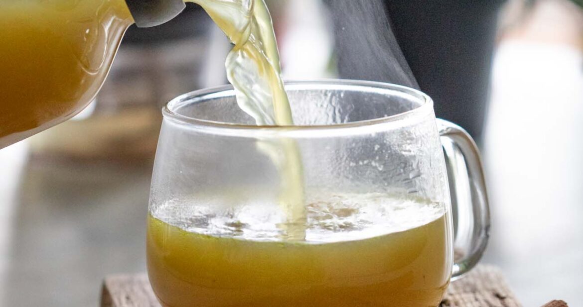 shot of turmeric chicken bone broth being poured into a clear glass mug resting on a wooden board