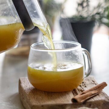 shot of turmeric chicken bone broth being poured into a clear glass mug resting on a wooden board