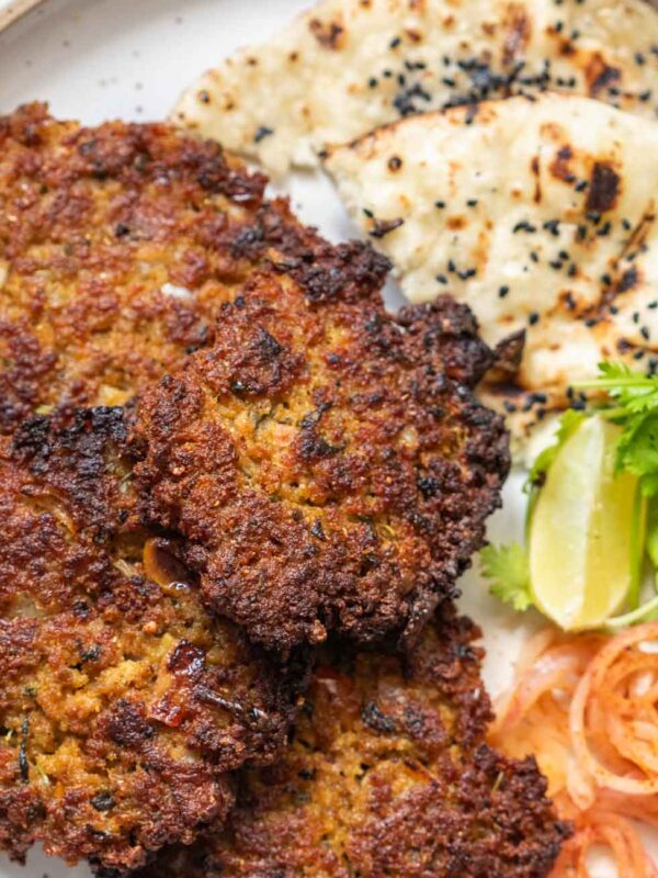 top shot of chapli kebabs served in a white plate with naan, onion, lemon, and coriamder