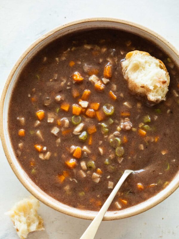 a whote bowl with ragi soup with diced carrots and beans with a spoon and a piece of bread dipped in the soup