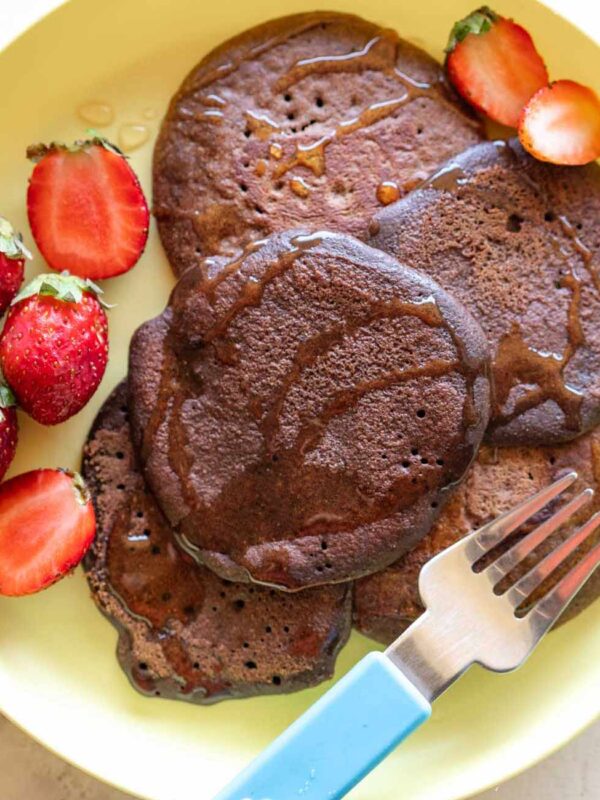 Chocolate Ragi Pancakes served on a yellow plate drizzled with honey and served alongside strawberries