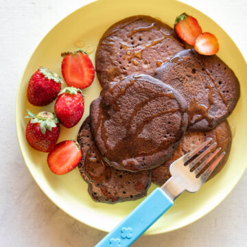 Chocolate Ragi Pancakes served on a yellow plate drizzled with honey and served alongside strawberries