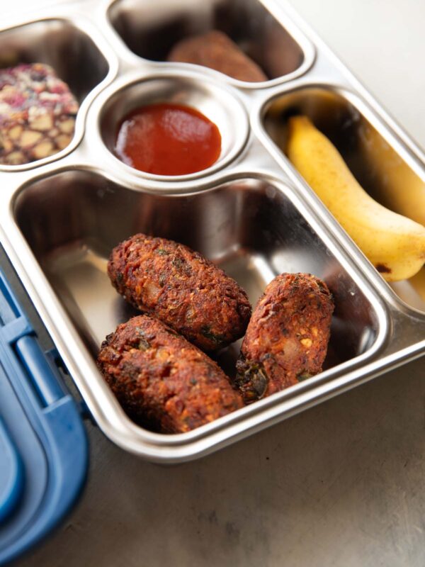 A picture of a lunch box with a sweet, ketchup, banana and a close up of the sprouts cutlet