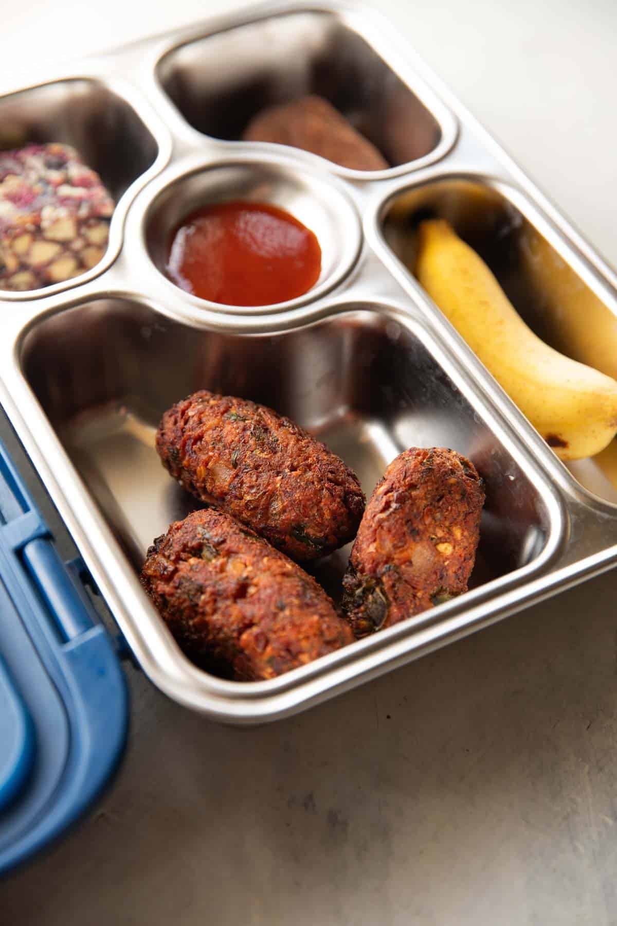 A picture of a lunch box with a sweet, ketchup, banana and a close up of the sprouts cutlet