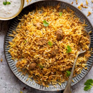 A plateful of mushroom biryani with a spoon on the side and a bowl of raita